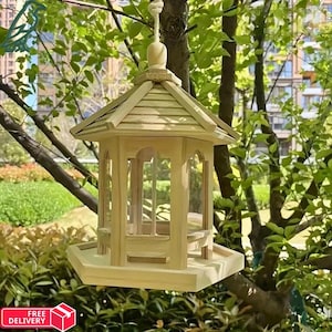 Könnte beinhalten: Ein Vogelhaus aus hellem Holz mit sechseckiger Basis und Satteldach. Das Futterhaus hat transparente Paneele und eine kleine Sitzstange. Es wird mit einem Seil an einem Ast aufgehängt. Die natürliche Holzfarbe fügt sich in das grüne Laub ein.