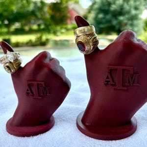 May include: Two maroon ring holders shaped like a hand with the letters "A" and "M" on the front. Each holder displays a gold ring. One ring has a large, clear stone, and the other has a red stone.