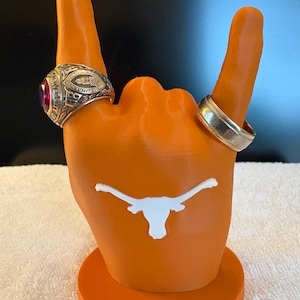 May include: Orange hand-shaped ring holder with a white longhorn logo. Two rings are displayed on the fingers. One ring has a red stone, and the other is silver. The base is round.