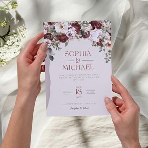 May include: A wedding invitation with floral accents. The invitation features the names "SOPHIA & MICHAEL" in a burgundy font, along with the date "OCT 18 2025". The invitation is held by two hands and is surrounded by a floral arrangement of burgundy and white flowers.