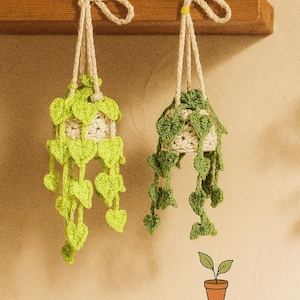 May include: Two handmade crochet plant hangers with leaf details. One is bright green, the other is a darker green. Both are suspended from a wooden shelf with natural rope. The image includes the text "Thee Littlest Plant Shoppe" and a plant pot graphic.