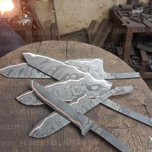 Peut inclure: Plusieurs lames de couteau non finies, en acier à motifs et avec des accents de cuivre, reposent sur une surface en bois. Les lames présentent un aspect stratifié distinctif, suggérant un processus de forgeage. L'arrière-plan montre un atelier avec des outils et un four lumineux.