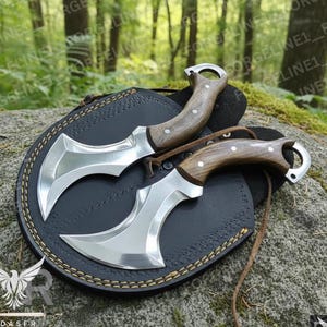 May include: Two silver and wood handled knives with curved blades rest on a black leather sheath. The handles are a light brown color. The sheath has a stitched border and a leather strap. The background is a forest setting.