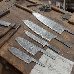 Peut inclure: Cinq lames de couteau en acier de Damas avec des motifs complexes et des accents cuivrés sont disposées sur un établi en bois. Les lames varient en taille et en forme, mettant en valeur une esthétique artisanale. L'atelier comprend des outils et des matériaux.