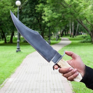 May include: A large, hand-forged knife with a curved, dark gray blade. The handle is off-white with a metal guard. The knife is held against a blurred outdoor background of a walking path and trees.
