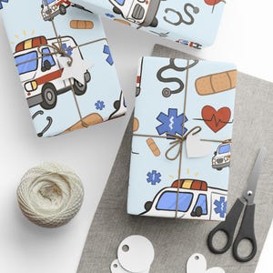 May include: Gift-wrapped presents with light blue paper featuring ambulances, stethoscopes, bandages, and hearts. The gifts are tied with twine and adorned with tags. A ball of twine, scissors, and blank tags are also visible.