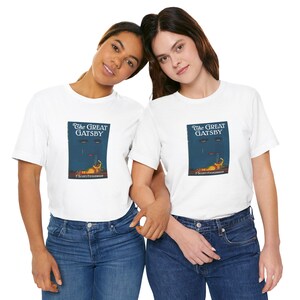 K&ouml;nnte beinhalten: Zwei Personen tragen wei&szlig;e T-Shirts mit einem Aufdruck des Buchcovers von "The Great Gatsby". Das Design befindet sich auf einem marineblauen Hintergrund mit dem Titel in Wei&szlig; und einer farbenfrohen Illustration. Sie tragen blaue Jeans.