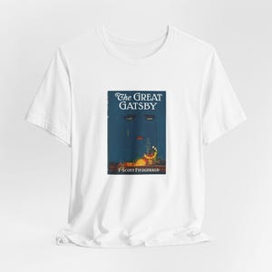 K&ouml;nnte beinhalten: Wei&szlig;es T-Shirt mit dem Cover von "The Great Gatsby" von F. Scott Fitzgerald. Das Design zeigt ein stilisiertes Gesicht mit Augen und einer roten Lippe, vor einem dunkelblauen Hintergrund. Der Titel ist in eleganter Schrift.