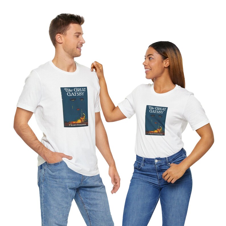 K&ouml;nnte beinhalten: Wei&szlig;e T-Shirts mit dem Cover von "The Great Gatsby" von F. Scott Fitzgerald. Das Design zeigt einen dunkelblauen Hintergrund mit orangefarbenen und roten Akzenten. Die Tr&auml;ger tragen blaue Jeans.