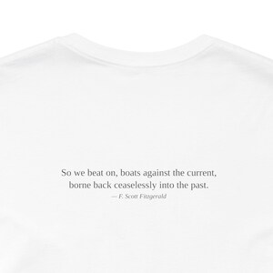 K&ouml;nnte beinhalten: Wei&szlig;es T-Shirt mit dem Zitat von F. Scott Fitzgerald: &bdquo;So we beat on, boats against the current, borne back ceaselessly into the past.&ldquo; Der Text ist in einer einfachen, eleganten Schriftart zentriert.