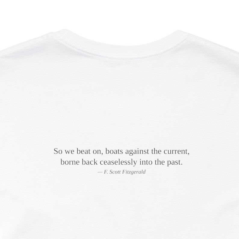 K&ouml;nnte beinhalten: Wei&szlig;es T-Shirt mit dem Zitat von F. Scott Fitzgerald: &bdquo;So we beat on, boats against the current, borne back ceaselessly into the past.&ldquo; Der Text ist in einer einfachen, eleganten Schriftart zentriert.