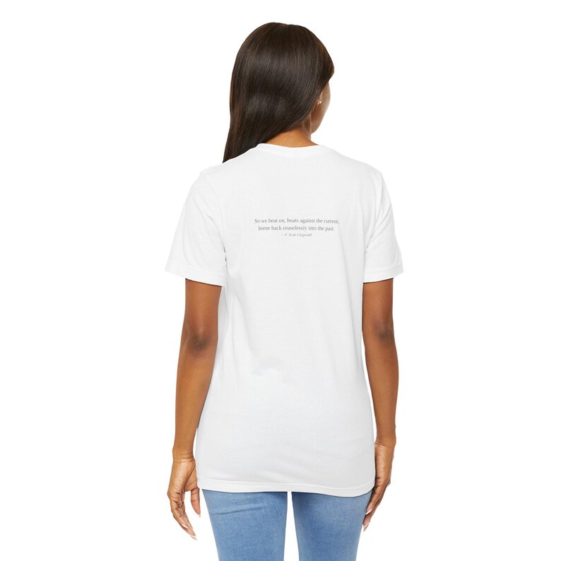 K&ouml;nnte beinhalten: Wei&szlig;es T-Shirt mit einem Zitat auf der R&uuml;ckseite. Das Zitat lautet: &bdquo;So we beat on, boats against the current, borne back ceaselessly into the past.&ldquo; - F. Scott Fitzgerald.
