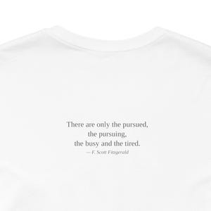 K&ouml;nnte beinhalten: Wei&szlig;es T-Shirt mit dem Zitat "There are only the pursued, the pursuing, the busy and the tired." von F. Scott Fitzgerald in Schwarz aufgedruckt. Das T-Shirt hat einen Rundhalsausschnitt und ist aus weichem Stoff gefertigt.