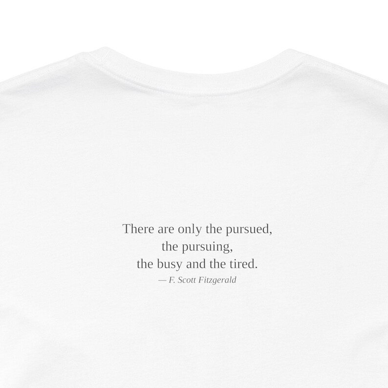 K&ouml;nnte beinhalten: Wei&szlig;es T-Shirt mit dem Zitat "There are only the pursued, the pursuing, the busy and the tired." von F. Scott Fitzgerald in Schwarz aufgedruckt. Das T-Shirt hat einen Rundhalsausschnitt und ist aus weichem Stoff gefertigt.