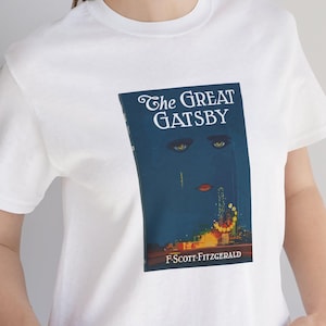 K&ouml;nnte beinhalten: Wei&szlig;es T-Shirt mit dem Cover von "The Great Gatsby" von F. Scott Fitzgerald. Das Design zeigt einen dunkelblauen Hintergrund mit stilisierten Augen, einer roten Lippe und einer Stadtlandschaft.