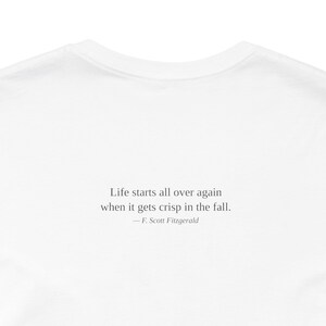 K&ouml;nnte beinhalten: Wei&szlig;es T-Shirt mit dem Zitat "Das Leben beginnt von vorn, wenn es im Herbst knusprig wird." von F. Scott Fitzgerald. Der Text ist in einer einfachen, eleganten Schriftart gehalten.