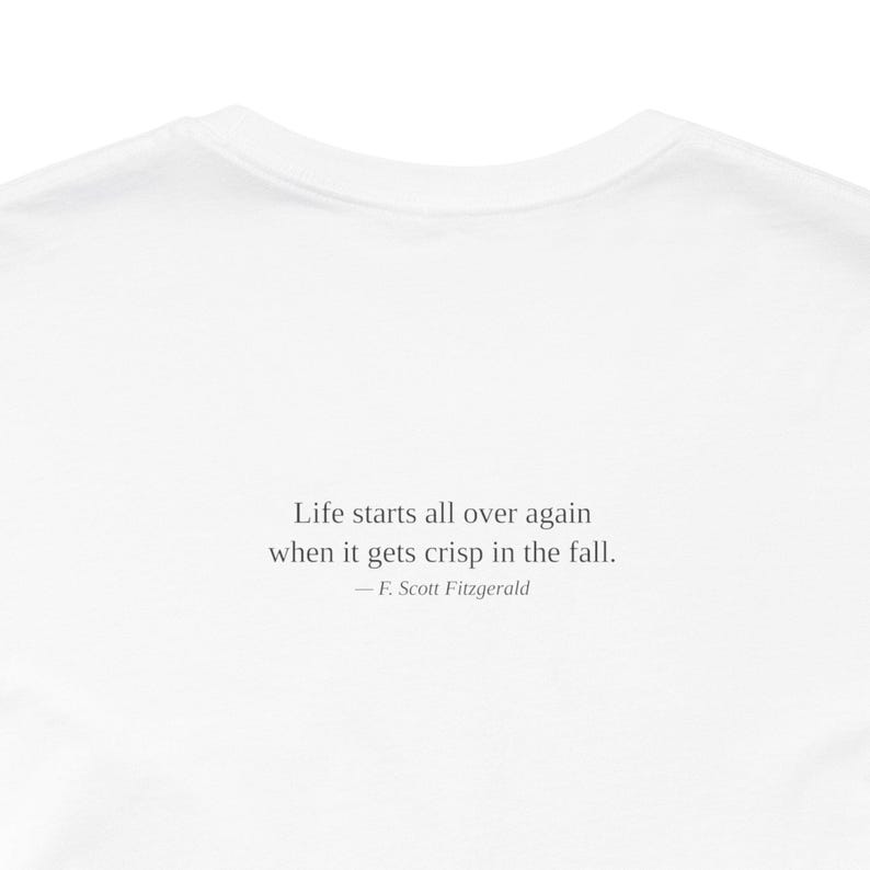 K&ouml;nnte beinhalten: Wei&szlig;es T-Shirt mit dem Zitat "Das Leben beginnt von vorn, wenn es im Herbst knusprig wird." von F. Scott Fitzgerald. Der Text ist in einer einfachen, eleganten Schriftart gehalten.