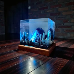 Könnte beinhalten: Ein klarer Kunstharzwürfel mit einer Miniatur-Stadtlandschaft und einer Godzilla-Figur, die einen blauen Strahl aussendet. Das Diorama steht auf einem Holzsockel, mit blauer Beleuchtung. Ein detailliertes, künstlerisches Display.