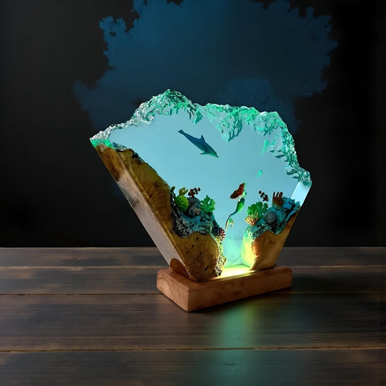 May include: A unique resin art piece depicting an underwater scene with a dolphin, mermaid, and coral. The artwork is encased in a clear, textured resin, mounted on a wooden base. The piece is illuminated with a soft, blue light.