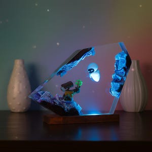 Könnte beinhalten: Eine transparente Resin-Skulptur mit den Charakteren Wall-E und Eve aus dem Animationsfilm. Die Skulptur hat einen Holzsockel und wird mit blauem Licht beleuchtet. Die Charaktere sind vor einer Kulisse aus blau beleuchteten Felsformationen platziert.