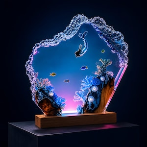 May include: A decorative resin sculpture featuring a mermaid swimming in a clear blue ocean scene with coral reefs and small fish. The piece is encased in a textured, wave-like resin frame and mounted on a rectangular wooden base with internal lighting.