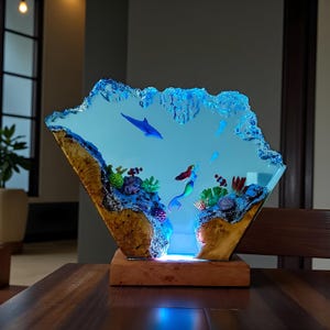 May include: A decorative resin sculpture featuring an underwater scene with a mermaid, fish, and coral. The sculpture has a blue and clear resin design with a wooden base. The overall shape is a diamond with a textured edge.