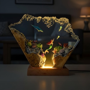May include: A decorative lamp featuring an underwater scene with a mermaid, dolphin, and jellyfish. The lamp is encased in clear resin with a wooden base and accents. The lamp is illuminated with a warm, inviting glow.