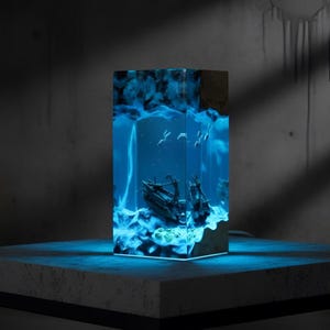 Può includere: Lampada rettangolare in resina con una scena di relitto illuminata da luce blu. La lampada presenta una parte superiore in legno e un diorama sottomarino dettagliato con una nave affondata ed elementi marini. La base emette una morbida luce blu.