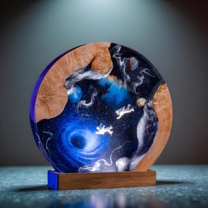 May include: A round, illuminated resin art piece depicting a cosmic scene with astronauts, nebulae, and a swirling galaxy. The artwork is framed by a wooden border and sits on a wooden base with a blue accent.
