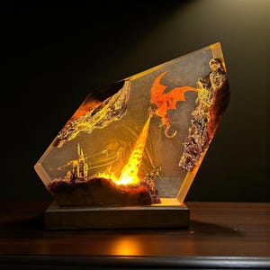 May include: A fantasy-themed resin lamp featuring a fiery orange dragon breathing fire towards a castle. The lamp has a wooden base and is encased in a clear, angled resin block with detailed rock formations and a warm, glowing light.