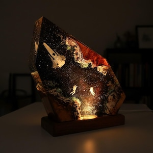 Könnte beinhalten: Dekorative Lampe mit einer Weltraum-Szene, eingebettet in einen transparenten, abgewinkelten Harzblock. Die Szene zeigt ein Raumschiff, Astronauten und einen Sternenhintergrund. Die Lampe steht auf einem dunklen Holzsockel und strahlt ein warmes Licht aus.