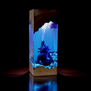 May include: A rectangular wooden block encases a blue resin scene featuring a Godzilla figure. The scene is illuminated with blue light, creating a dramatic effect. The top of the block has a dark, textured finish.