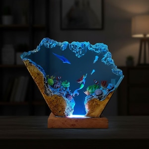 May include: A decorative resin lamp featuring an underwater scene with a mermaid, fish, and coral. The lamp has a wooden base and a blue, textured top, creating a glowing effect. The lamp is approximately 8 inches tall.