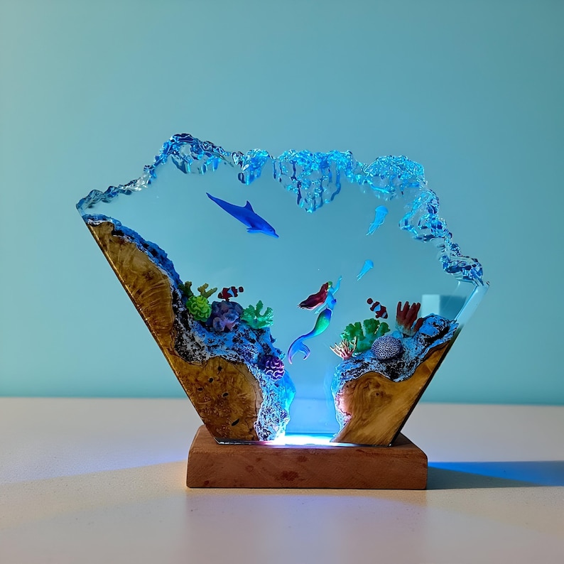 May include: A decorative resin sculpture featuring an underwater scene with a mermaid, dolphin, and coral. The sculpture is set on a wooden base and has a blue and clear color scheme. The design includes a light that illuminates the scene.