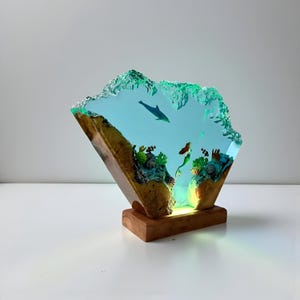 May include: A decorative resin sculpture depicting an underwater scene with a dolphin, mermaid, and coral. The sculpture is illuminated with a soft green light and set on a wooden base. The resin is clear with a turquoise hue.