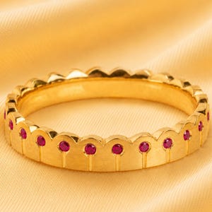 Puede incluir: Un anillo de oro con un diseño de borde festoneado en la parte superior. El anillo está engastado con pequeñas piedras preciosas redondas de color rojo rosado vibrante, incrustadas en la base de cada festón, creando un patrón repetitivo.