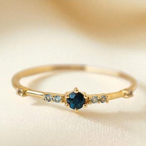 Puede incluir: Un delicado anillo de oro con una piedra preciosa azul oscuro central, flanqueada por piedras más pequeñas de color azul claro. El anillo tiene una banda fina y un diseño simple y elegante. Las piedras preciosas están engastadas en un engaste de garras. Es una joya.