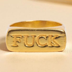 May include: A gold-toned metal ring featuring a rectangular face with the word FUCK embossed in capital letters. The ring has a smooth, polished finish and a thick band design, showcasing a bold, metallic aesthetic with clear, legible lettering.