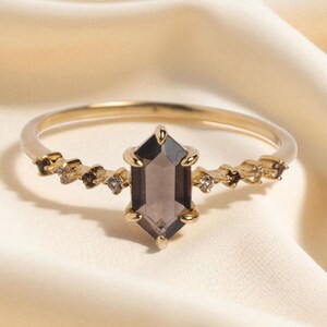 May include: A gold ring with a hexagonal smoky quartz gemstone at its centre. The ring has a slender band and is embellished with small, sparkling details on either side of the main stone. The ring is set against a soft, cream-coloured background.