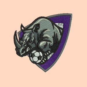 May include: Embroidered patch featuring a gray rhinoceros with a soccer ball, set against a purple and white shield. The rhino is detailed with shading, giving it a 3D effect. The soccer ball is black and white.