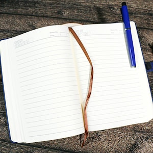 Puede incluir: Un cuaderno abierto con una cubierta azul y un marcador de cinta marr&oacute;n. Un bol&iacute;grafo azul descansa sobre la p&aacute;gina derecha. El cuaderno est&aacute; sobre una superficie de madera, con una pi&ntilde;a y un peque&ntilde;o objeto azul cerca.