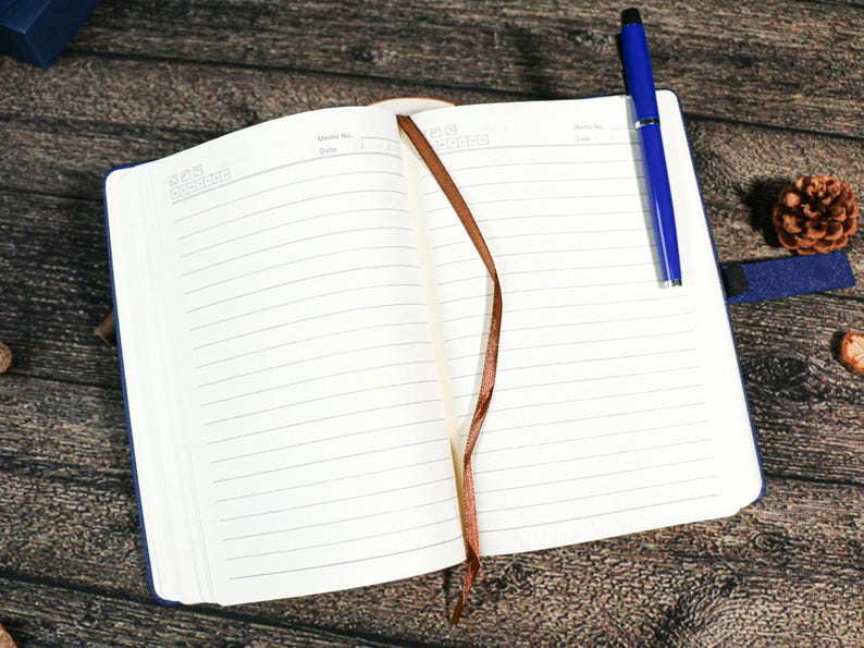 Puede incluir: Un cuaderno abierto con una cubierta azul y un marcador de cinta marr&oacute;n. Un bol&iacute;grafo azul descansa sobre la p&aacute;gina derecha. El cuaderno est&aacute; sobre una superficie de madera, con una pi&ntilde;a y un peque&ntilde;o objeto azul cerca.