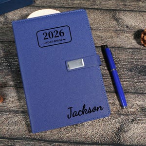 Puede incluir: Una agenda azul de cuero de 2026, un d&iacute;a por p&aacute;gina, con un cierre plateado y un bol&iacute;grafo azul a juego. El nombre "Jackson" est&aacute; impreso en la portada. El fondo es una superficie de madera con elementos decorativos.