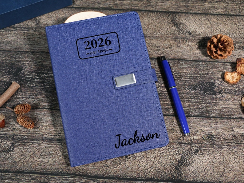 Puede incluir: Una agenda azul de cuero de 2026, un d&iacute;a por p&aacute;gina, con un cierre plateado y un bol&iacute;grafo azul a juego. El nombre "Jackson" est&aacute; impreso en la portada. El fondo es una superficie de madera con elementos decorativos.
