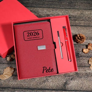 Puede incluir: Un juego de diario y bol&iacute;grafo de polipiel roja en una caja de regalo roja. El diario est&aacute; grabado con "2026 -DAY APAGE-" y el nombre "Pete". El juego incluye un bol&iacute;grafo rojo y un bol&iacute;grafo plateado. La caja est&aacute; abierta.