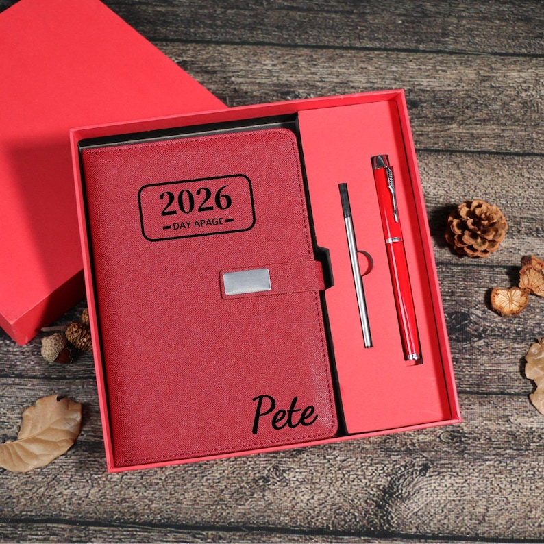 Puede incluir: Un juego de diario y bol&iacute;grafo de polipiel roja en una caja de regalo roja. El diario est&aacute; grabado con "2026 -DAY APAGE-" y el nombre "Pete". El juego incluye un bol&iacute;grafo rojo y un bol&iacute;grafo plateado. La caja est&aacute; abierta.