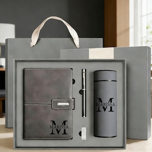Op de afbeelding: Een grijze geschenkset met een notitieboek, pen, waterfles en een kleine metalen clip. Het notitieboek en de waterfles zijn gepersonaliseerd met de naam "MARISSA". De set wordt gepresenteerd in een grijze geschenkdoos met een bijpassende geschenkzak.