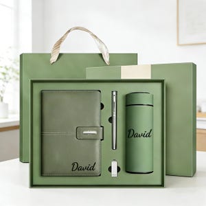 Puede incluir: Un juego de regalo verde que contiene un cuaderno, un bolígrafo y un vaso, todos con el nombre "David" impreso. El juego se presenta en una caja verde con una bolsa de regalo a juego. El cuaderno tiene un cierre.