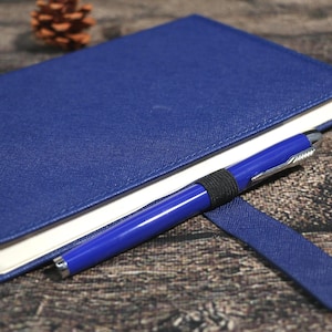Puede incluir: Un cuaderno azul con un bol&iacute;grafo a juego. El cuaderno tiene una cubierta texturizada y p&aacute;ginas con bordes blancos. El bol&iacute;grafo azul, con detalles plateados, se sujeta con una banda el&aacute;stica negra.