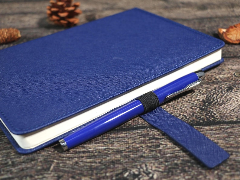 Puede incluir: Un cuaderno azul con un bol&iacute;grafo a juego. El cuaderno tiene una cubierta texturizada y p&aacute;ginas con bordes blancos. El bol&iacute;grafo azul, con detalles plateados, se sujeta con una banda el&aacute;stica negra.